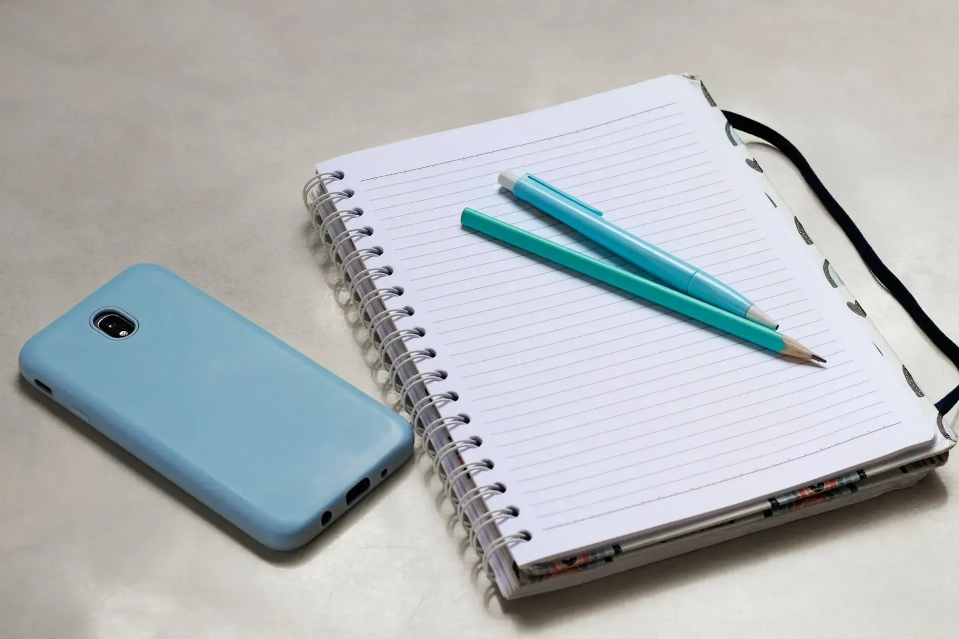 Cuaderno con espiral acompañado de un lápiz, un bolígrafo y un móvil sobre una superficie clara.
