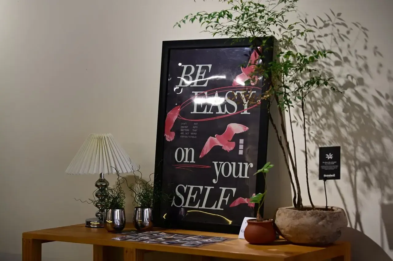 Póster motivacional enmarcado con mensaje tipográfico y elementos gráficos, acompañado de plantas y una lámpara sobre una mesa.
